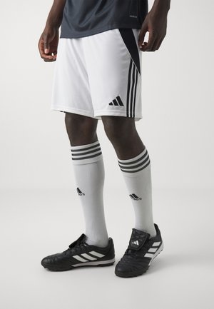 Mężczyzna w białych i czarnych szortach piłkarskich Adidas, białych wysokich skarpetach Adidas i czarnych korkach Adidas, stojący na jednolitym tle.