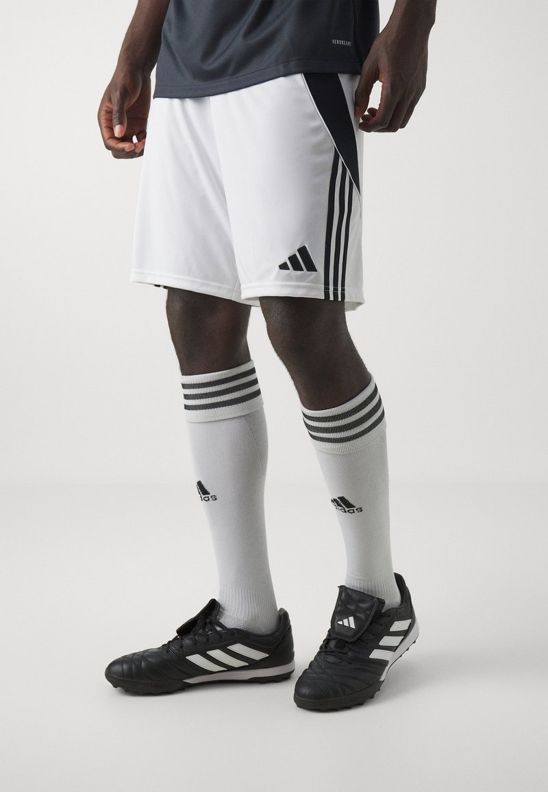 Homme portant un short de soccer Adidas blanc et noir, des chaussettes montantes blanches Adidas, et des crampons noirs Adidas, debout sur un fond uni.