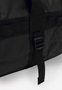 Sac de voyage noir en tissu texturé, avec une bandoulière réglable et une fermeture en plastique. Design simple et fonctionnel sans accents supplémentaires.