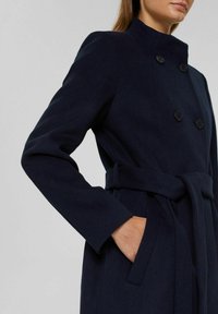 Marineblauwe dubbelzijdige jas van zacht materiaal, met een hoge kraag, lange mouwen, zijzakken en een strikriem in de taille.