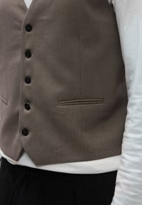 Καφέ υφασμάτινο γιλέκο με έξι μαύρα κουμπιά, πλαϊνές τσέπες και λεία υφή. Φορεμένο πάνω από λευκό πουκάμισο με μακριά μανίκια και μαύρο παντελόνι.