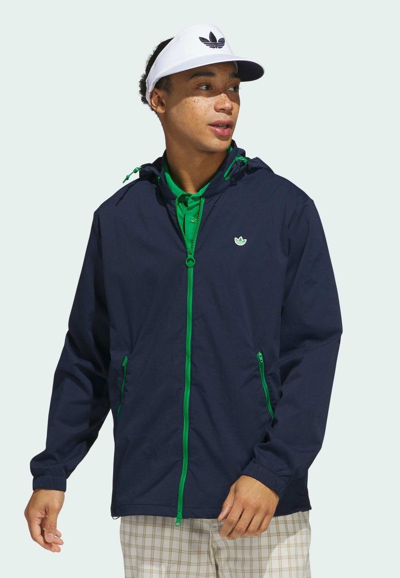 Chaqueta cortavientos de la marina con acentos en verde en la cremallera, capucha ajustable y bolsillos laterales. Se lleva sobre una camisa verde de cuello y pantalones de cuadros en color beige.