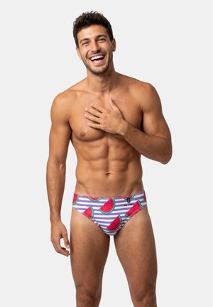 Homme souriant et en forme portant un slip de bain coloré à motif pastèque sur fond blanc uni, main touchant la poitrine.