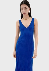 Robe bleue à côtes, sans manches, avec un décolleté en V profond, présentant une silhouette ajustée et une longueur maxi. Texture lisse avec une légère brillance.