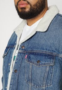 Džinsinis švarkas su šviesios spalvos dirbtinio kailio apykakle, mėlynos spalvos plaunamas, auksinėmis siūlėmis ir dviem krūtinės kišenėmis. Levi's logotipas matomas šone.