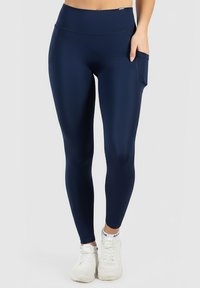 Tmavě modré legíny vyrobené z elastického materiálu, s vysokým páskem a boční kapsou. Nucená s bílými sportovními botami.