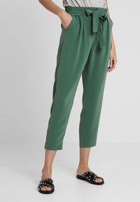 Pantaloni verdi a vita alta con cintura a fiocco, gambe affusolate, tasche laterali e una consistenza liscia. Abbinati a sandali neri con borchie.