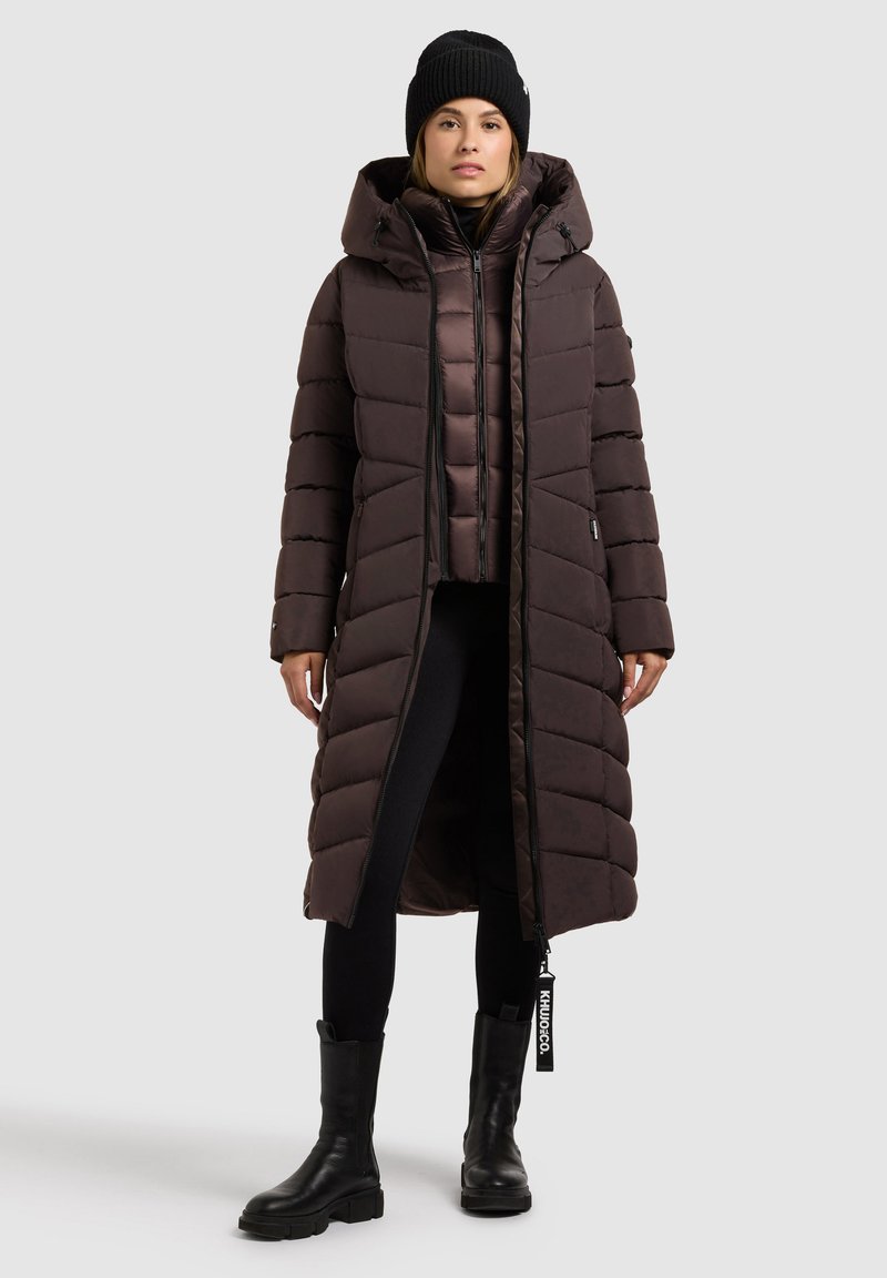 Langer brauner Daunenmantel mit Kapuze, mit horizontalen Steppnähten, einem Reißverschluss vorne und Taschen. Getragen über einem schwarzen Oberteil und Leggings, dazu schwarze Stiefel.