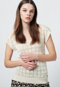 Stickad topp i krämfärg med korta ärmar, som har ett öppenstickat mönster av rutor. Ribbad nederkant och sidoknappdetalj. Slät, lätt textur.