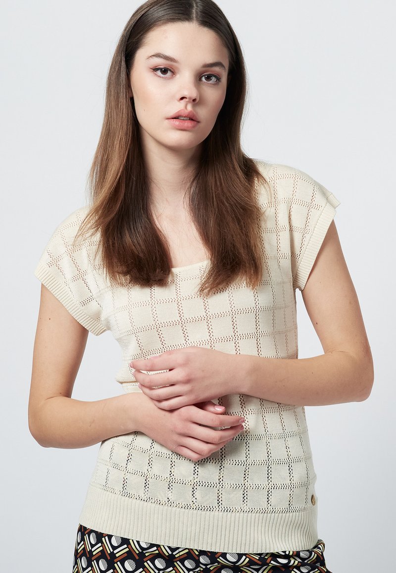 Stickad topp i krämfärg med korta ärmar, som har ett öppenstickat mönster av rutor. Ribbad nederkant och sidoknappdetalj. Slät, lätt textur.