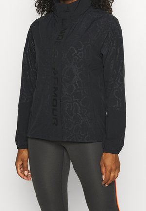 Donna che indossa una giacca nera a fantasia Under Armour con collo alto e leggings neri con una striscia arancione sul lato.