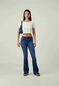 Ung kvinne som står, iført en hvit cropped knappebluse, blå slengjeans, svarte platåsko, og bærer en mørkeblå skulderveske.