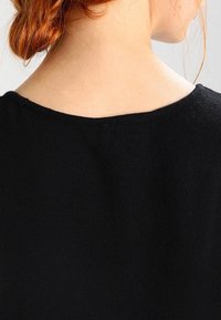 Svart stickad tröja med rundad halsringning. Har en slät textur och subtila sömmar längs kanterna. Nära vy bakifrån.