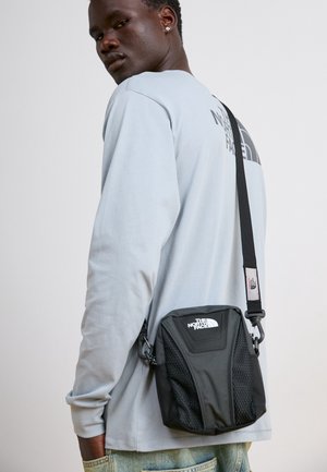 Jeune homme portant une chemise à manches longues bleu clair et un sac bandoulière noir The North Face, regardant par-dessus son épaule sur un fond uni.