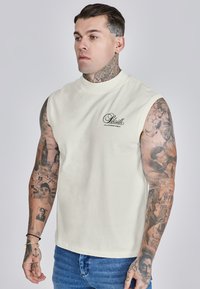 Camiseta sin mangas de algodón blanca con escote redondo y un pequeño logotipo negro en el pecho, combinada con unos vaqueros de mezclilla azul.