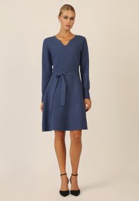 Abito lavorato a maglia blu con scollo a V, maniche lunghe e cintura annodata in vita. Orlo svasato e texture liscia. Indossato con tacchi neri.