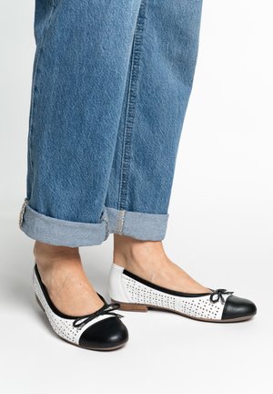 Pieds portant des ballerines perforées blanches et noires avec des nœuds, associées à un jean en denim bleu retroussé sur fond blanc.