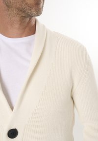 Kootud kreemjast villast kardigan, millel on šaalikaelus, üks must nupp ja tekstureeritud soonik. Kantud valge T-särgi peal.