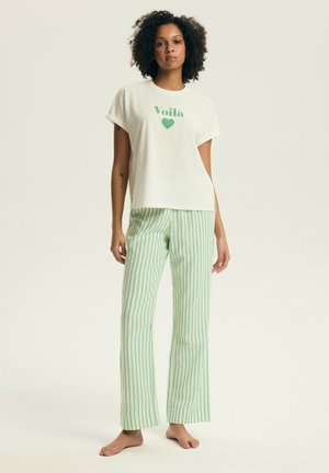 Kobieta o kręconych włosach stoi boso, ubrana w białą koszulkę z napisem "Voilà" i sercem oraz jasne zielone spodnie w paski.