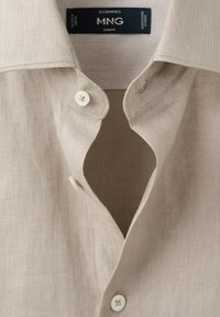 Lysebeige hørskjorte med krave, knapdetalje og MNG Essentials-mærke indeni halsudskæringen.
