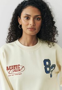 Kremowy sweatshirt z czerwonym haftowanym napisem "Pacific Coast City by the Bay" oraz niebieską kraciastą cyfrą "87" z przodu.
