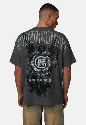 Mężczyzna o krótkich włosach, noszący ciemnoszarą, oversize'ową koszulkę z grafiką anioła oraz tekstem "Established 2011 British Atelier" i "North West, England".