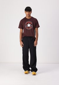 T-shirt graphique marron arborant un logo circulaire "Converse All Star", associé à un pantalon noir ample et des baskets avec des accents jaunes.