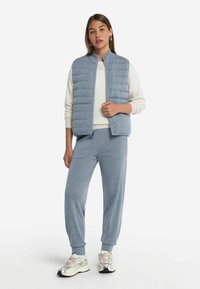 Ljusblå vadderad väst med hög krage, vit långärmad tröja och matchande blå joggarbyxor. Silverfärgade sportskor med detaljer.