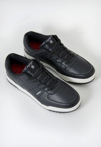 Adidași din piele neagră cu detalii perforate, talpă din cauciuc alb și șireturi negre. Prezintă interior roșu și branding pe limbă.