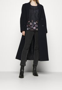 Mujer vestida con un abrigo largo negro sobre una blusa oscura con estampado, jeans negros y botines negros de tobillo, de pie contra un fondo blanco.