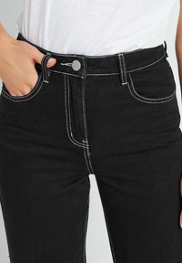 Jeans de mezclilla negra con costuras blancas en contraste, cintura alta y diseño de cinco bolsillos. Incluye un cierre de botón y trabillas para cinturón.