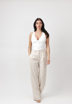 Mujer con cabello largo y oscuro, vestida con un top blanco sin mangas y pantalones beige de pierna ancha, de pie con una mano en el bolsillo contra un fondo liso.