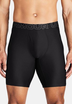 Męski tors w czarnych krótkich spodenkach kompresyjnych z paskiem "UNDER ARMOUR", stojący z ramionami rozluźnionymi na białym tle.