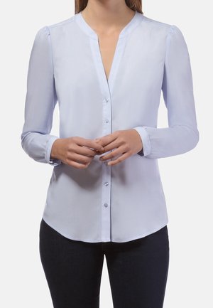 Blusa de manga larga azul claro con botones, usada por una persona con las manos entrelazadas al frente, combinada con pantalones oscuros.