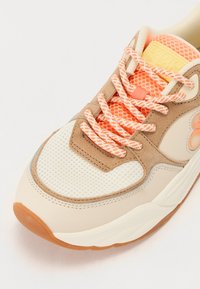 Beige sneaker featuring a mesh toe, tan accents, peach laces with a checkered pattern, and a rubber sole. Logo on tongue in yellow.