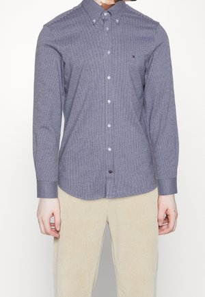 Man wearing a fitted blue and white vertical striped button-up shirt with beige corduroy pants against a white background.