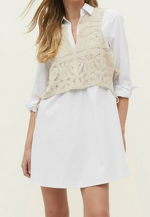 Femme portant une robe chemise blanche à manches longues et un gilet sans manches en crochet beige noué sur les côtés.
