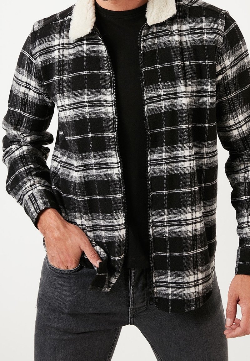 Schwarz-weiß kariertes Jackett mit durchgehendem Reißverschluss und Kunstpelzkragen. Lange Ärmel, strukturierter Stoff, über einem schwarzen Hemd getragen.