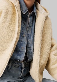 Kremowa polarowa kurtka o miękkiej fakturze, z zamkiem błyskawicznym i kapturem. Noszona na dżinsowej koszuli i spódnicach z wysokim stanem.