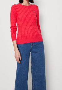 Woman wearing a bright pink cable-knit sweater and blue wide-leg jeans, standing against a plain light background.
