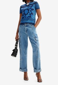 Top de punto azul con un patrón y mangas cortas, con un motivo central de sol; combinado con unos vaqueros cargo azul claro con múltiples bolsillos.