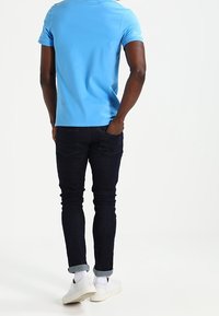 Ljusblå skjorta med kort ärm, mörka denimjeans, vänd mot bakvyn. Vita sneakers med uppvikta byxben. Slätt tyg, figursydd design.