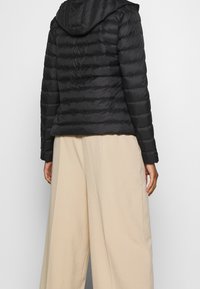 Černá péřová bunda s kapucí, prošívaný design a elastické manžety, kombinovaná s širokými béžovými kalhotami. Matný povrch, lehký materiál.