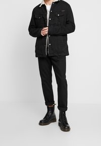 Svart denimjacka med sherpa-foder, två framfickor och knappstängningar, kombinerad med svarta byxor och svarta snörstövlar.