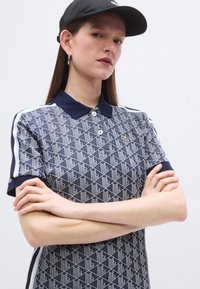 A tengerészkék mintás pólógeometrikus mintákkal, egyszínű gallérral, két gombbal és oldalt fehér csíkokkal; a modell hozzáillő fekete sapkát visel.