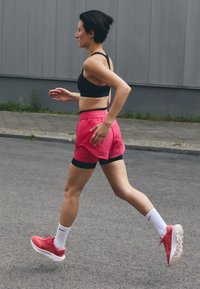 Løper ikledd en svart sports-bh, rosa shorts og røde joggesko. Merkbare detaljer inkluderer hvite sokker og en glatt, atletisk kroppsbygning.