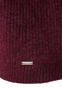 Burgunderfarbener Strickstoff mit einem angenähten Metallschild, das mit "MORE & MORE" in schwarzen Buchstaben graviert ist.