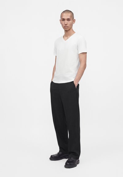 Homme debout, les mains dans les poches, portant un t-shirt blanc à manches courtes et col en V, un pantalon noir et des chaussures noires sur un fond uni.