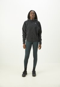 Czarna polarowa kurtka z zamkiem i kapturem, ciemnoniebieskie legginsy oraz czarne buty sportowe. Zawiera logo Nike na kurtce i legginsach.
