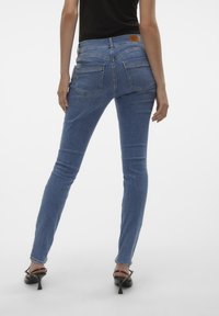 Jeans ajustados de color azul con bolsillos traseros, fabricados en tela de mezclilla. Presentan una etiqueta de cuero marrón en la cintura. Se combinan con tacones negros.